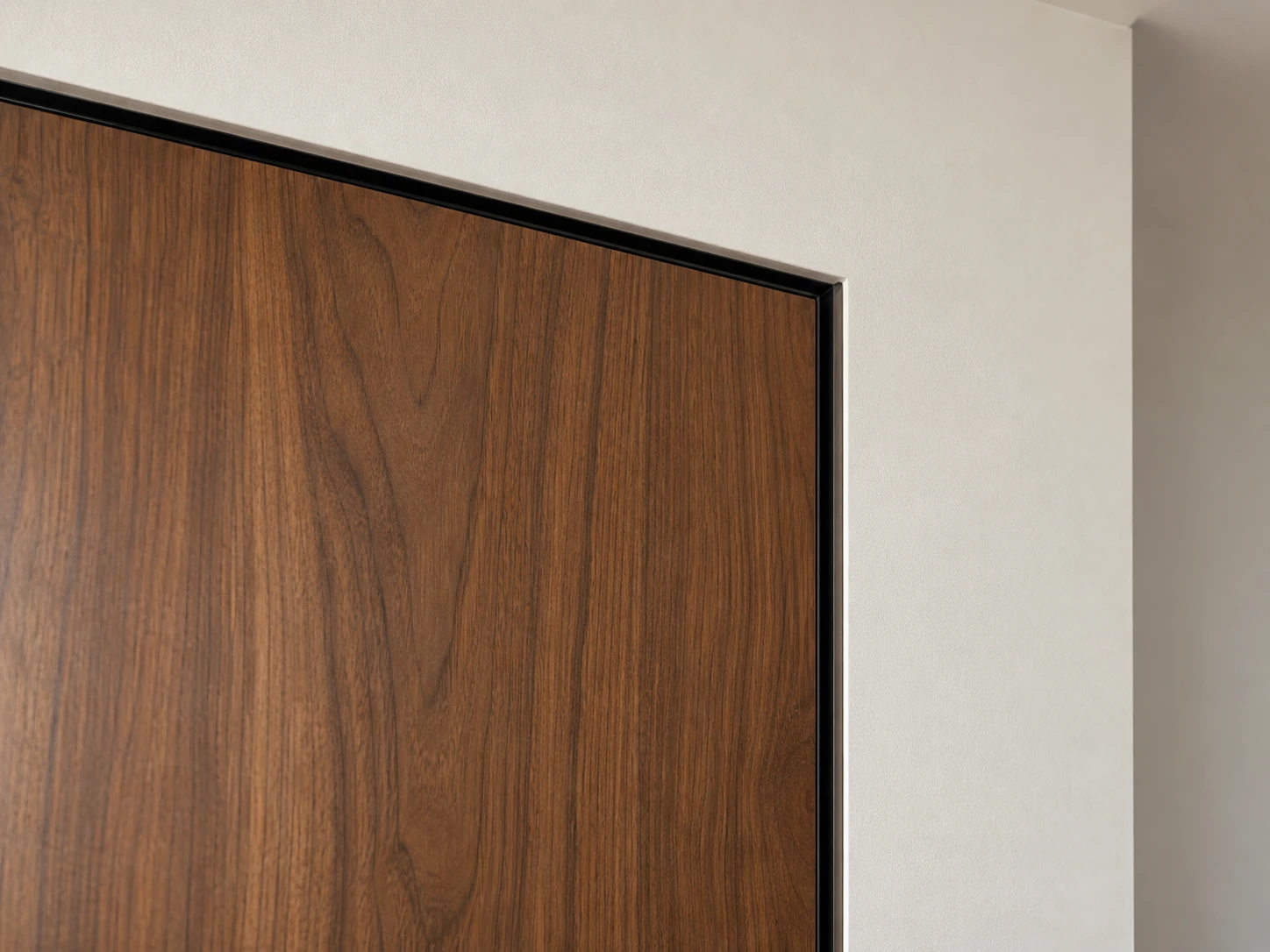 Narrow recessed trim framing the corner of a walnut door against a finished drywall wall.